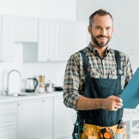 smiling handsome plumber holding clipboard and looking at camera in kitchen