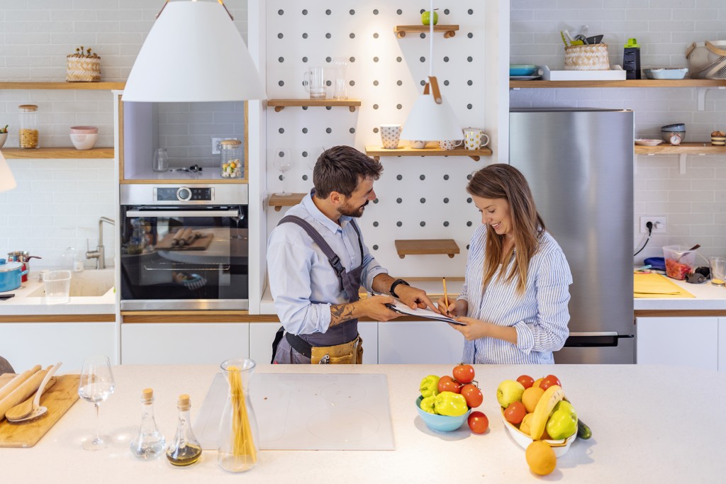 Happy Woman Writing On Clipboard In Front Of Young Plumber Standing In Kitchen, signing Invoice From Male Plumber In Kitchen