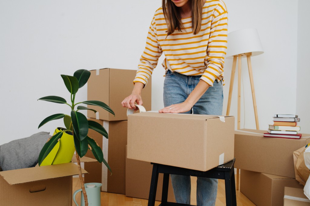 Middle age woman in casual clothes standing next to pile of boxes, packing, she's moving out from an old apartment. Sealing cardboard boxes with adhesive scotch tape. Close up, cropped, no head.