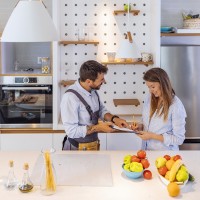 Happy Woman Writing On Clipboard In Front Of Young Plumber Standing In Kitchen, signing Invoice From Male Plumber In Kitchen