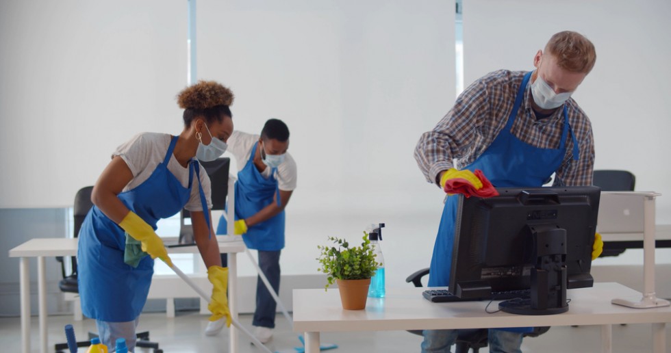 Team of diverse young cleaning service professionals in safety mask at work in office. Multiethnic janitors wearing protective mask and uniform cleaning modern office