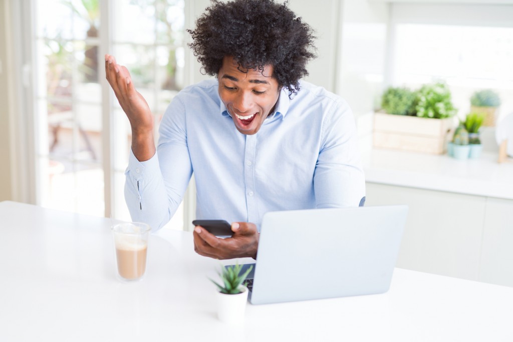 African American business man using smartphone and laptop very happy and excited, winner expression celebrating victory screaming with big smile and raised hands