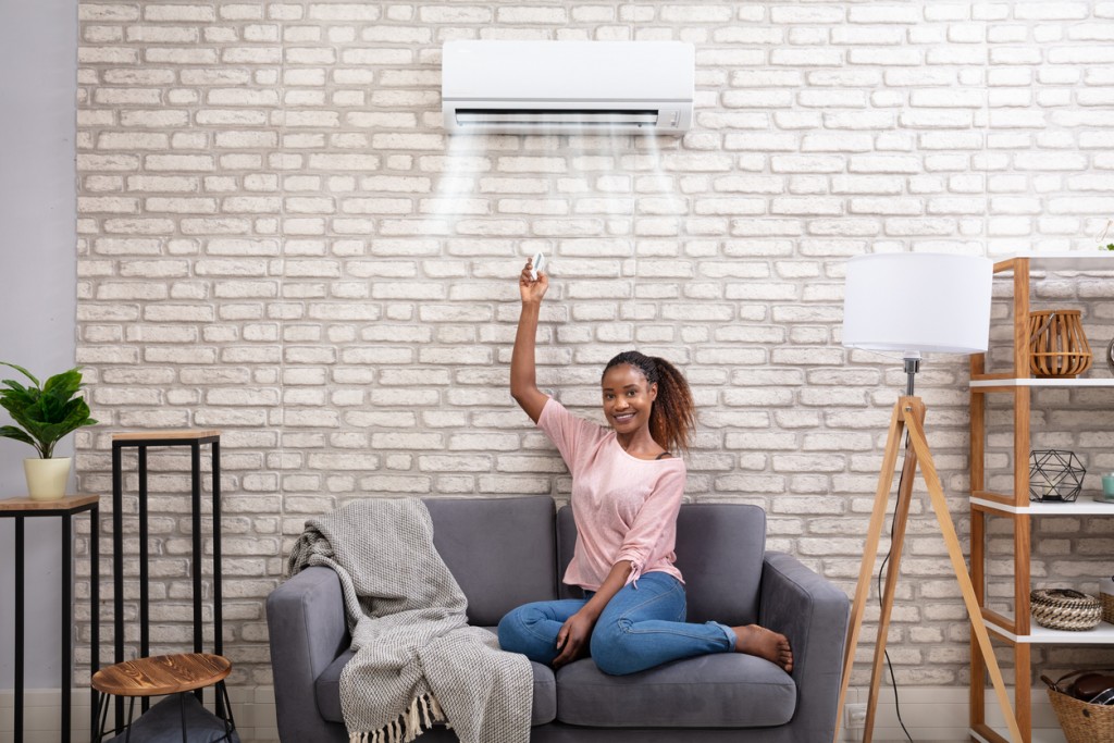 Young African Woman Switching On Air Conditioner With Remote Control At Home