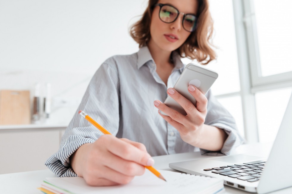 serious-young-woman-glasses-holding-mobile-phone-taking-notes-while-sitting-light-apartment